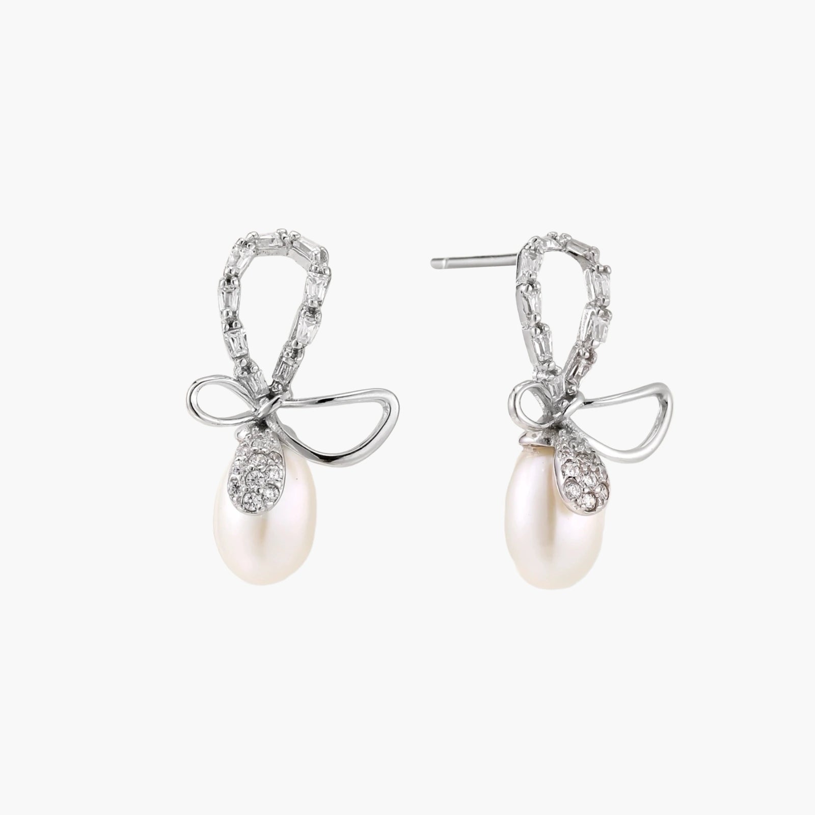 Silver bow-shaped stud earrings with baguette crystals and dangling white freshwater pearls on cream background