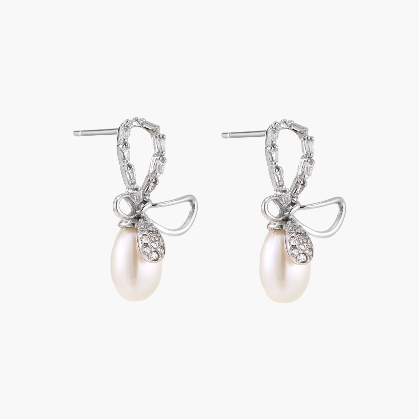 Silver bow-shaped stud earrings with baguette crystals and dangling white freshwater pearls on cream background