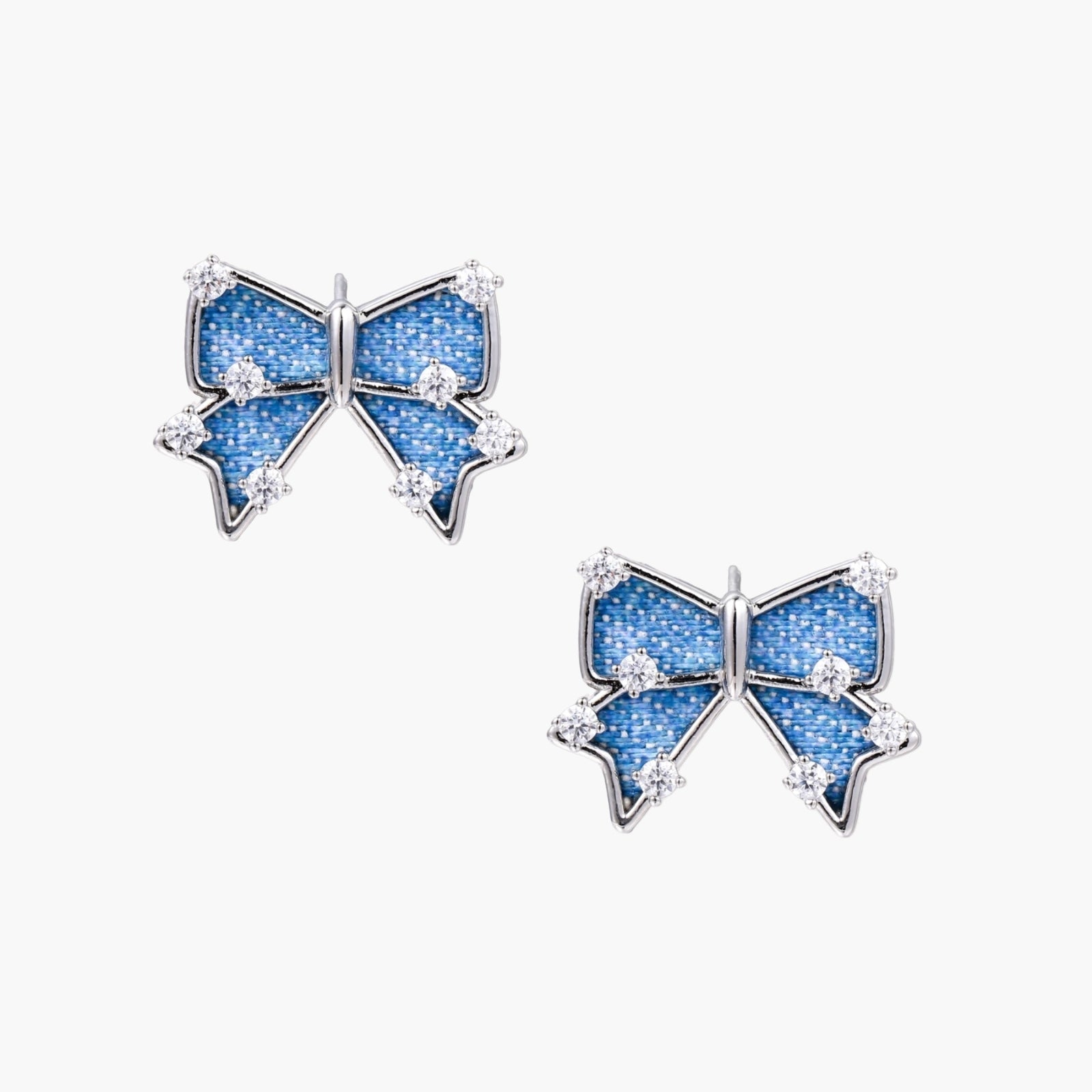 Blue denim bow stud earrings with white gold plating and crystal accents on cream background