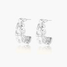 White gold plated sculptural hoop earrings with organic wavy edges and reflective metallic finish on cream background