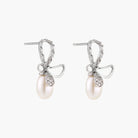 Silver bow-shaped stud earrings with baguette crystals and dangling white freshwater pearls on cream background