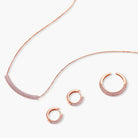 Rose gold jewelry set with pavé cubic zirconia stones including curved necklace, hoop earrings, and adjustable ring on cream background.